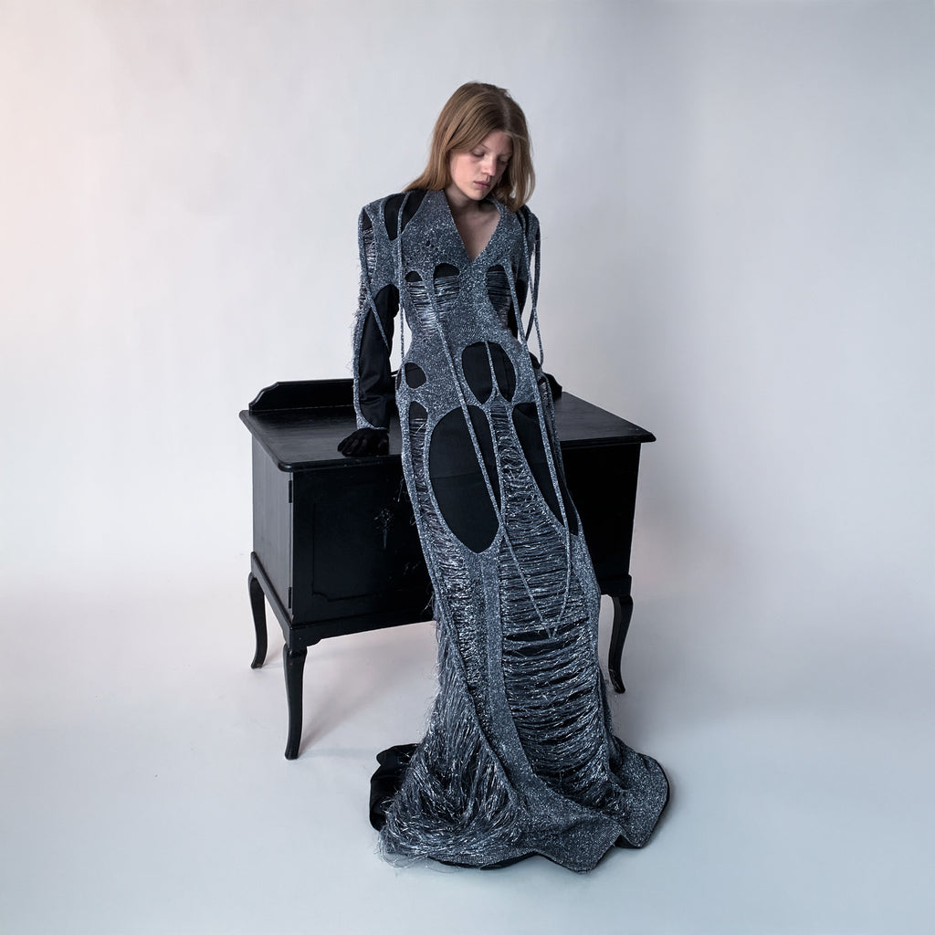 Woman wearing a long, dark gray dress layered with metallic knit standing next to a black desk.