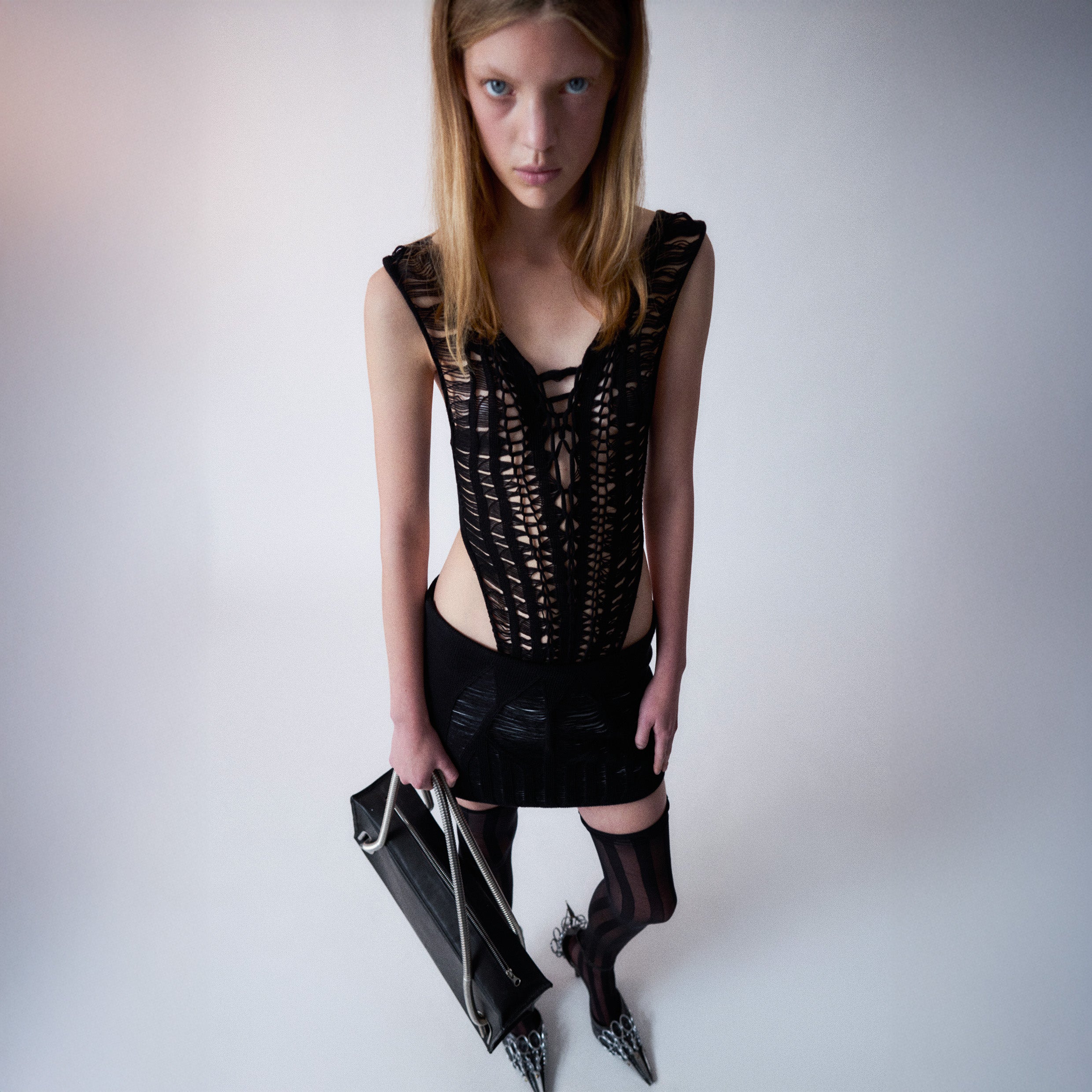 Person Wearing a black knitted top and a black knitted skirt, holding a black leather bag with metallic handles on a light gray background 