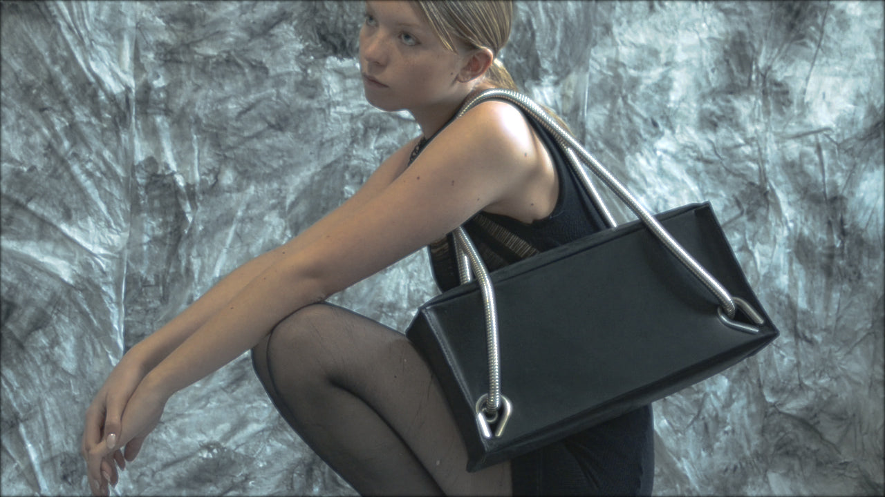 Person in a black knitted dress wearing a bllack leather handbag with silver handles and on a textured background 