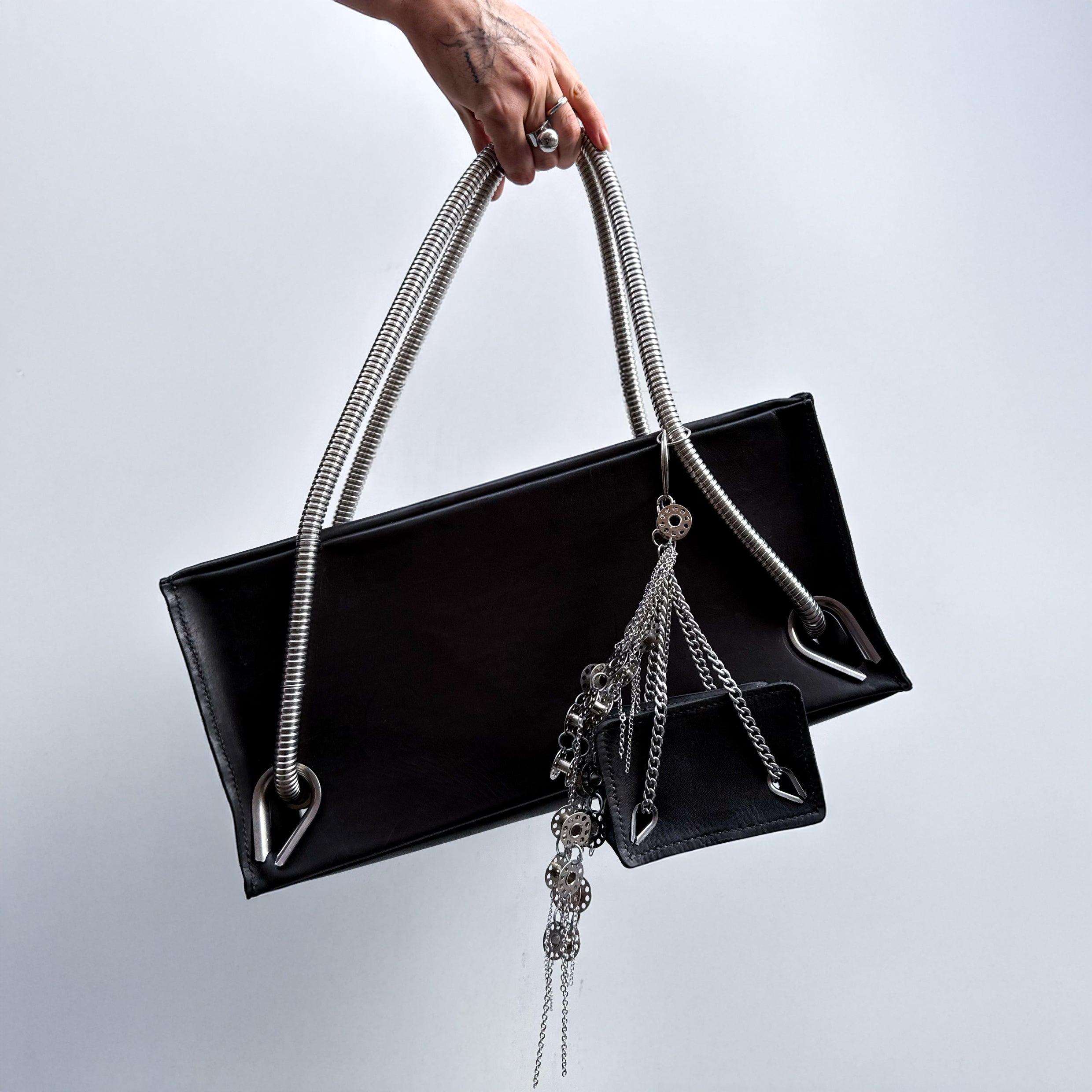 Black leather handbag with silver handles with  a mini black leather bag with metal chain handles as a bag charm and  a keychain with metal bobbins and chains